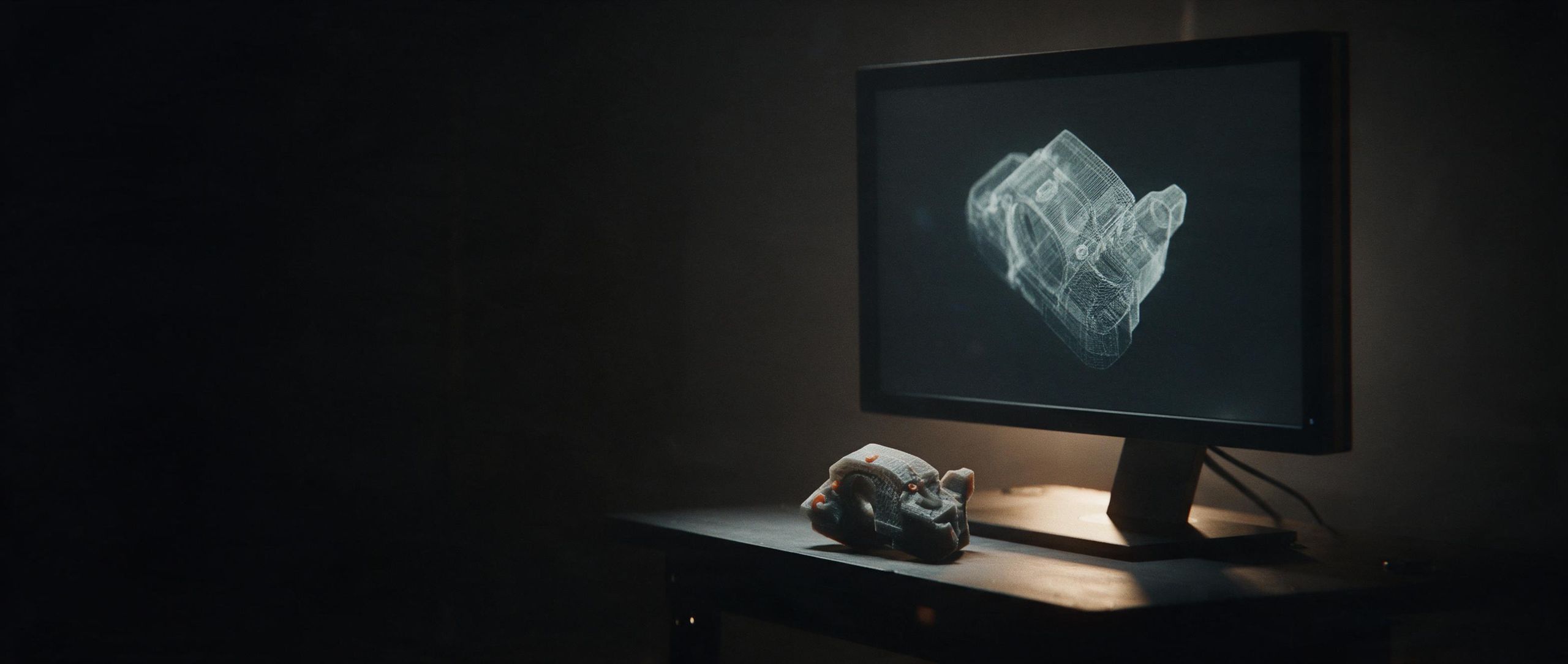 Una pieza impresa en 3D sobre un escritorio, delante de un monitor con su modelo de alambre.