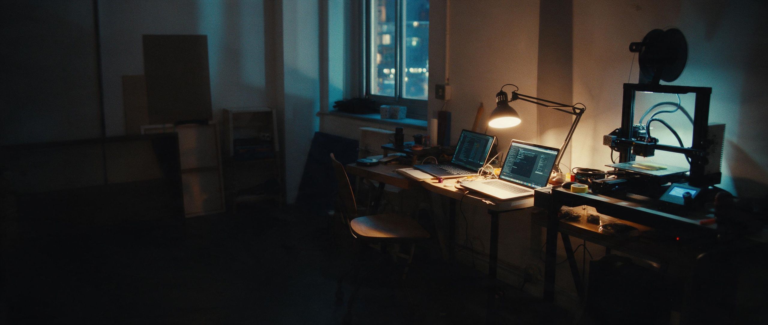 Two desks in a small studio at night, one lamp on, a printer working in the corner.